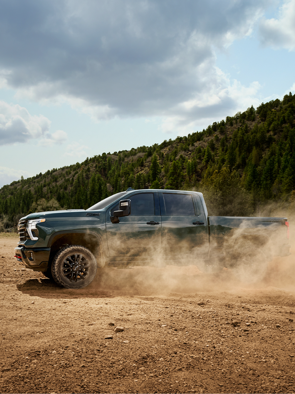 A Blue Chevy Silverado LD Driving Through a Dusty Dirt Road Surrounded by Tall Green Pine Trees and Mountains in the Distance.