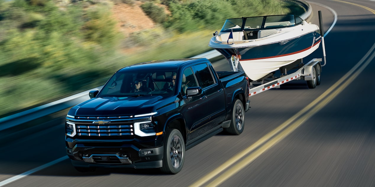 A 2027 Chevy Silverado 1500 Towing a Boat and Driving Around the Curve of a Road Next to a Lake