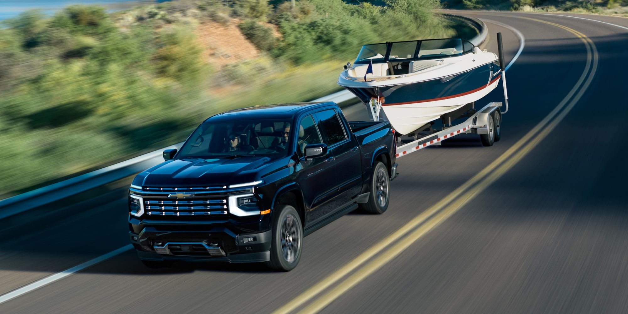 A 2027 Chevy Silverado 1500 Towing a Boat and Driving Around the Curve of a Road Next to a Lake