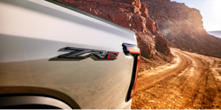 Three Quarters View of a White 2026 Chevy Colorado Driving through a Dust Cloud on a Desert