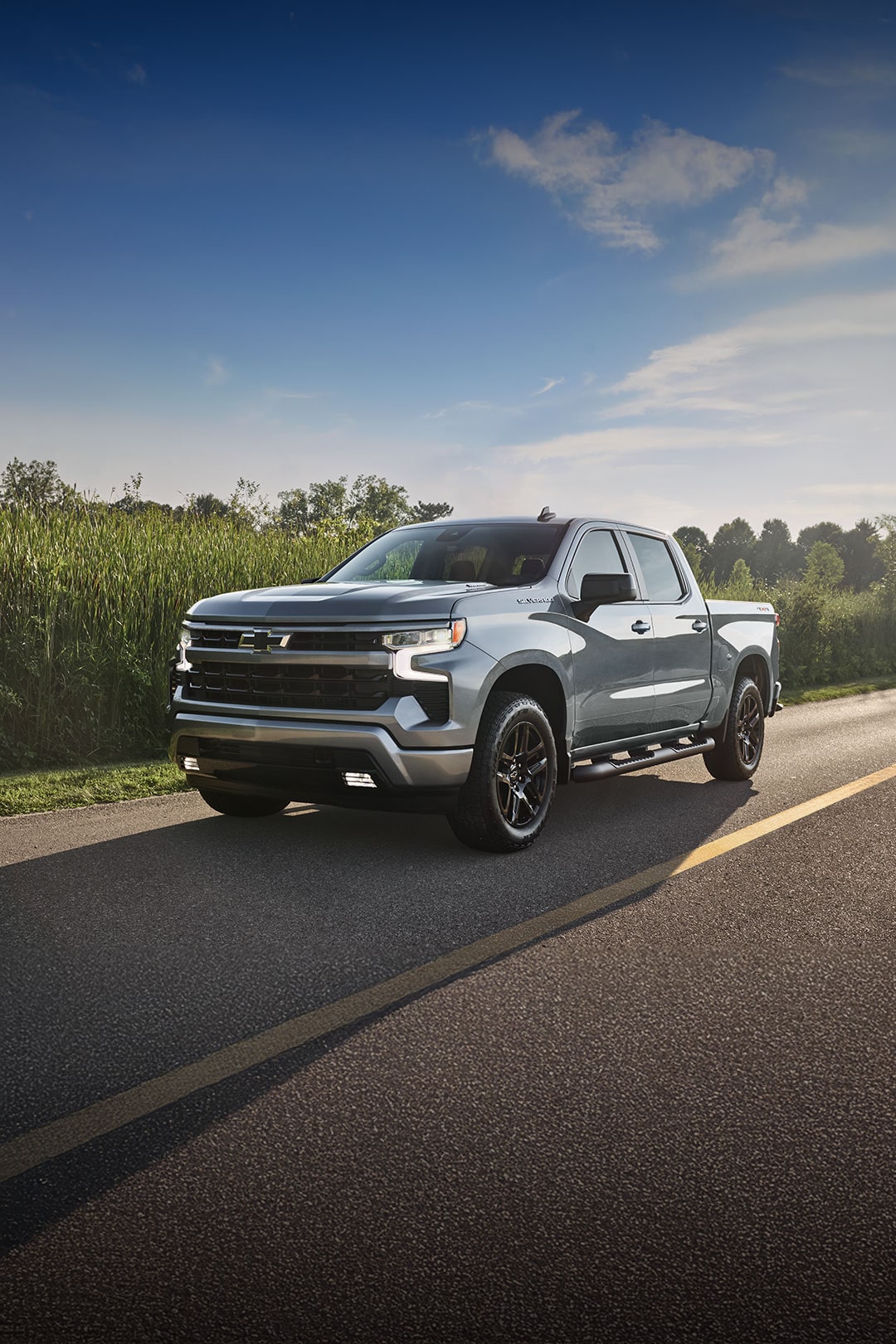 Three-quarter View of a Chevy Silverado 1500 Driving Down a Road Lined with Green Foliage with Text Overlay Reading "Can't wait?"