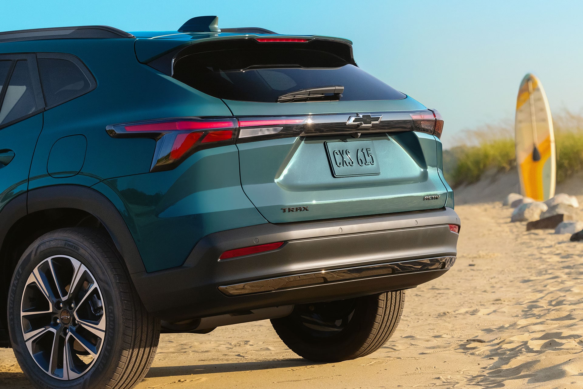 A Rear Three-Quarter View of a Chevy Trax Parked on a Sandy Beach With a Surfboard Standing Nearby.