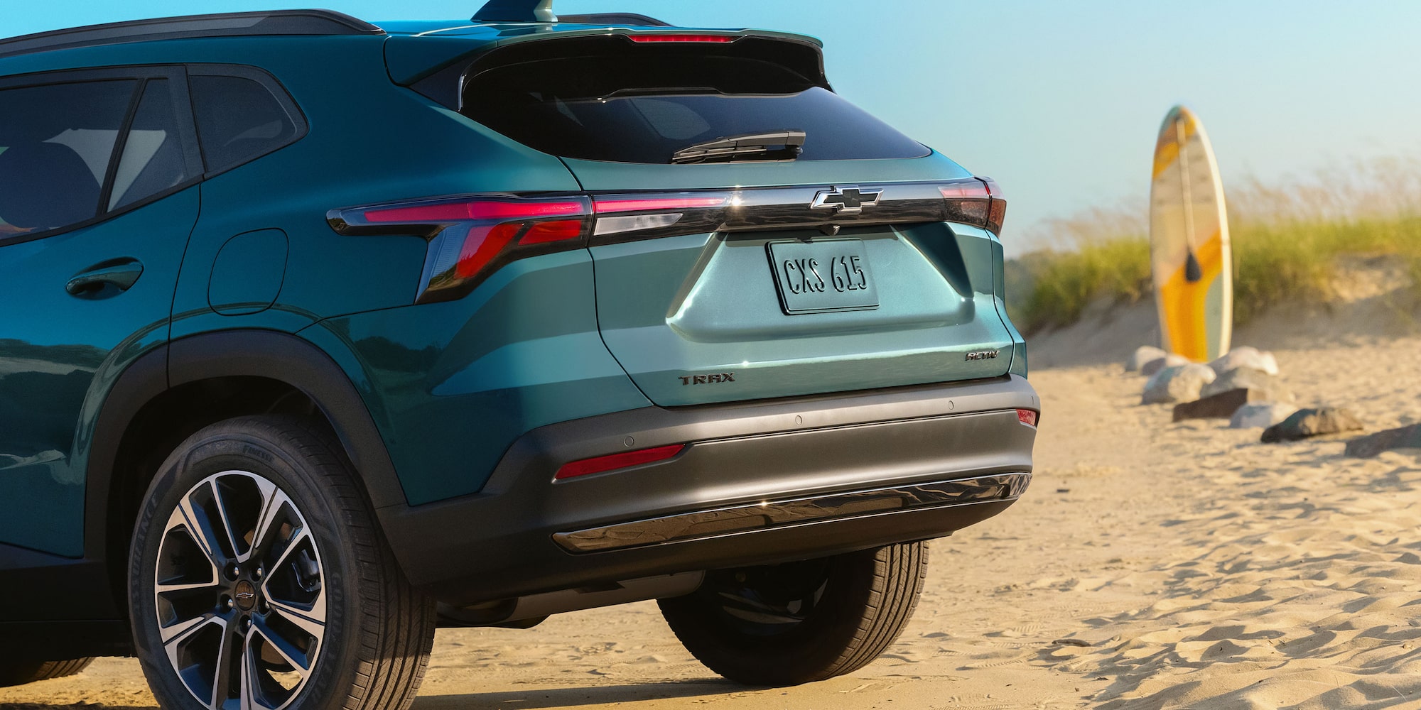 A Rear Three-Quarter View of a Chevy Trax Parked on a Beach With a Surfboard Standing Nearby.