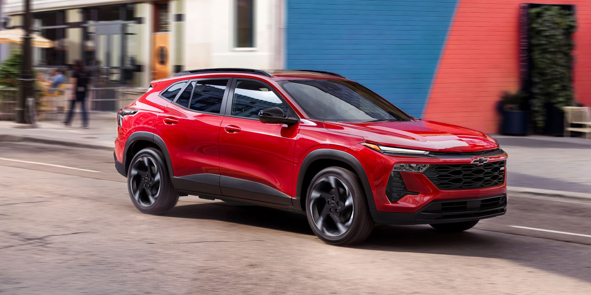 A Red Chevy Trax Driving Through a City Street Surrounded by Modern Buildings and Pedestrians.