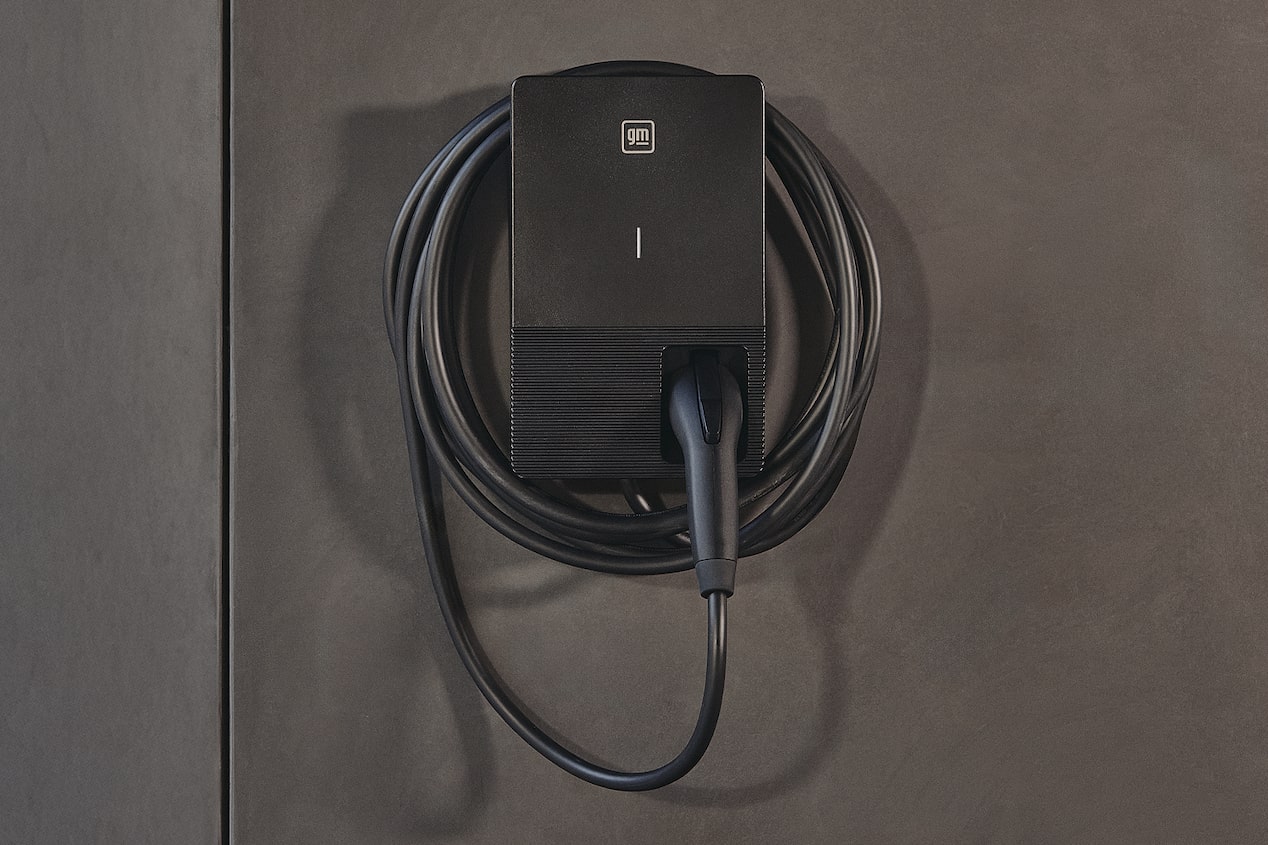 Un cargador GM PowerUp 2 montado en la pared con un cable de carga enrollado, asegurado prolijamente contra una superficie exterior gris lisa.