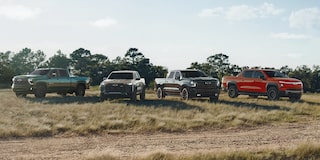 Four Chevrolet Pickup Trucks Including a Silverado ZR2, Silverado HD ZR2, and Colorado ZR2 Parked on a Dirt Road in a Grassy Field with Trees in the Background Under a Partly Cloudy Sky.