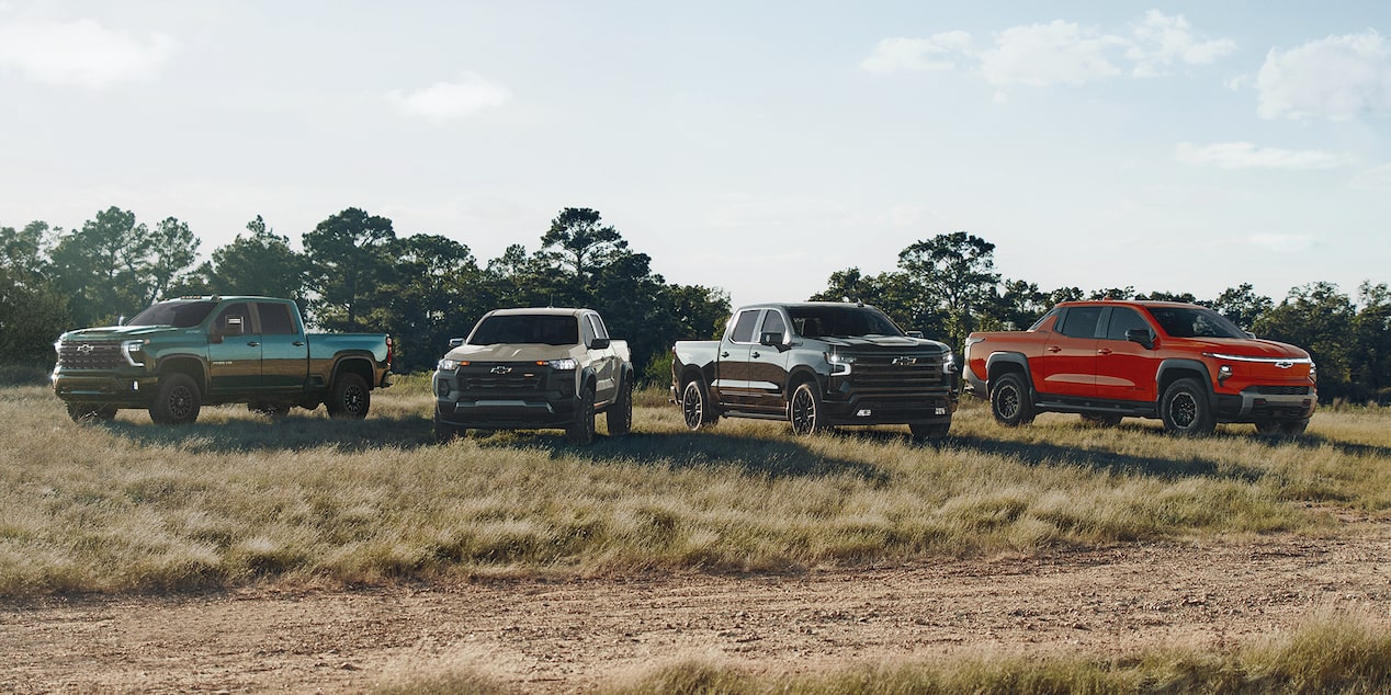 Four Chevrolet Pickup Trucks Including a Silverado ZR2, Silverado HD ZR2, and Colorado ZR2 Parked on a Dirt Road in a Grassy Field with Trees in the Background Under a Partly Cloudy Sky.