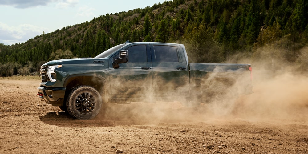Dark green pickup truck driving on a dirt road, kicking up dust, with forested hills in the background.