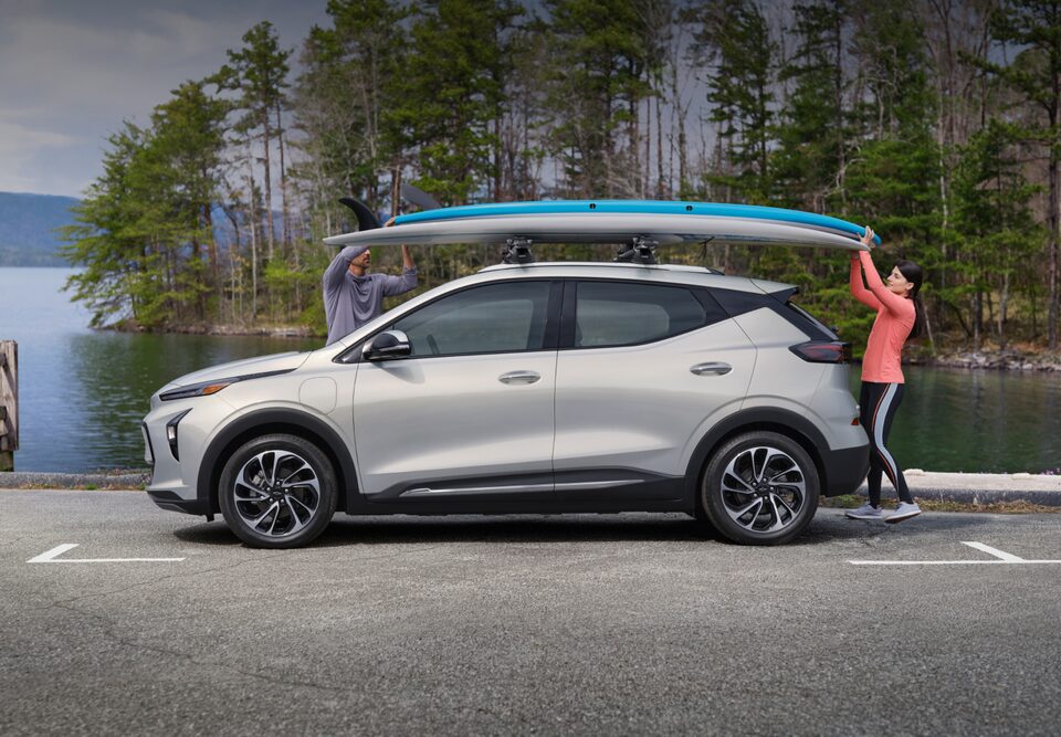 2023 Bolt EUV Parked in Parking Lot with Paddle Board on Roof and 2 People Reaching Up to Unload It