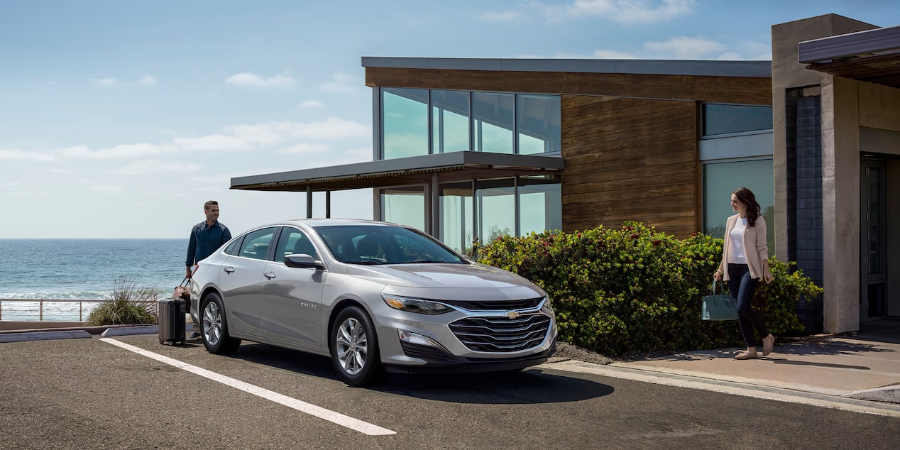 Side-Profile Of The 2024 Chevy Malibu In Sterling Gray Metallic Parked In Front Of Modern Building