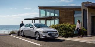 Side-Profile Of The 2024 Chevy Malibu In Sterling Gray Metallic Parked In Front Of Modern Building