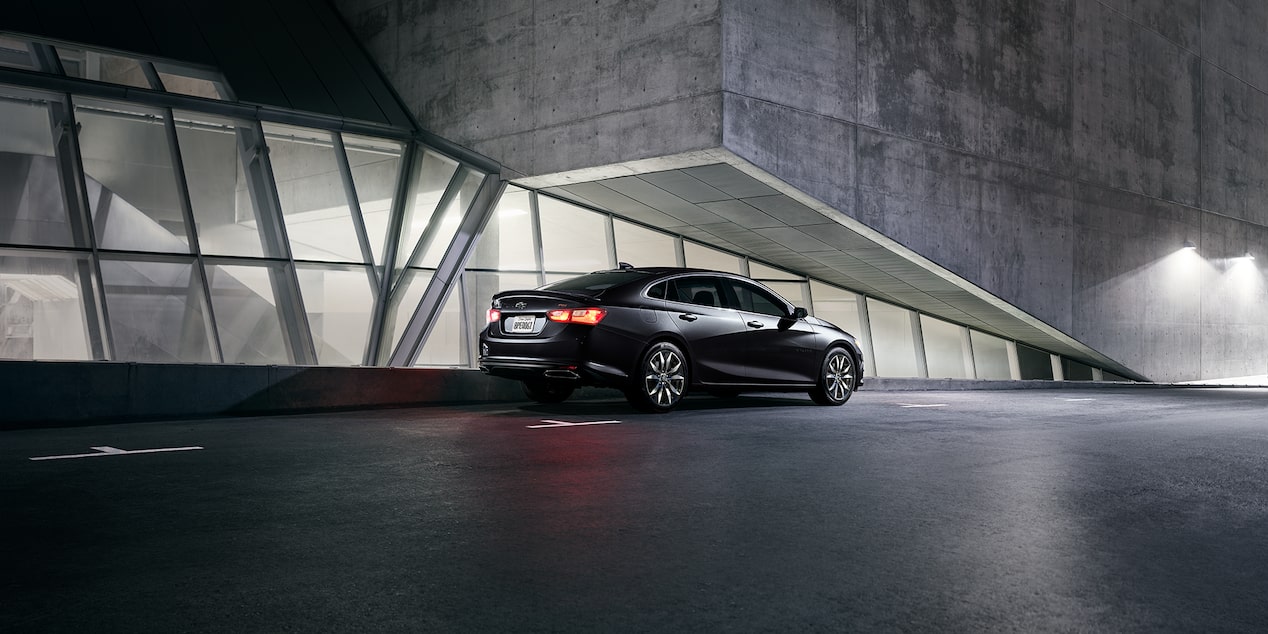 Side-Profile Of The 2024 Chevy Malibu In Mosaic Black Metallic Parked In Front Of Building Windows