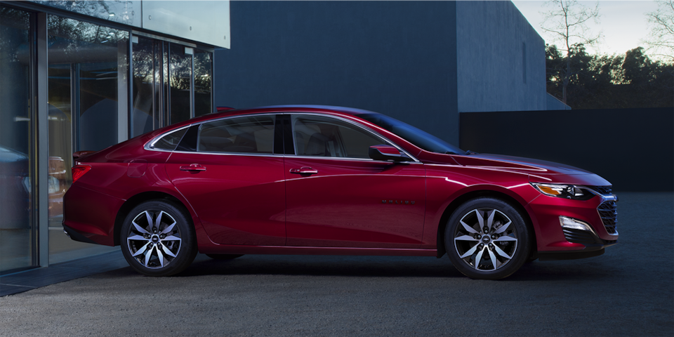 Side Profile Of The 2024 Chevy Malibu In Radiant Red Tintcoat Parked In Front Of A Modern Building
