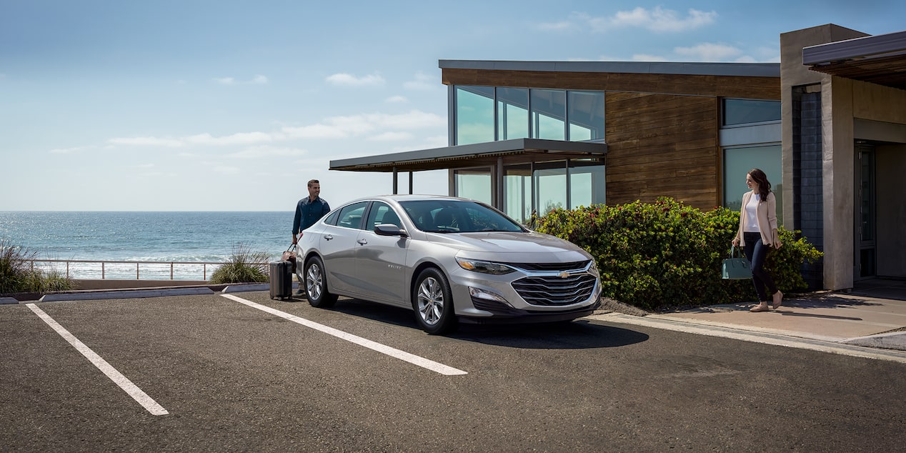 Una Chevy Malibu 2025 color Gris Esterlino estacionada frente a una casa bajo un cielo azul despejado