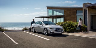 Una Chevy Malibu 2025 color Gris Esterlino estacionada frente a una casa bajo un cielo azul despejado