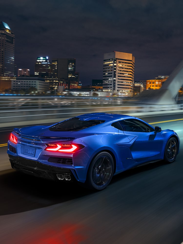Three Quarters Rear View of a Blue Chevy Corvette E-Ray Driving Down the Road Surrounded by Trees