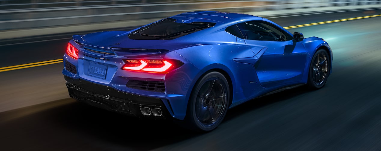 Three Quarters Rear View of a Blue Chevy Corvette E-Ray Driving Down the Road Surrounded by Trees