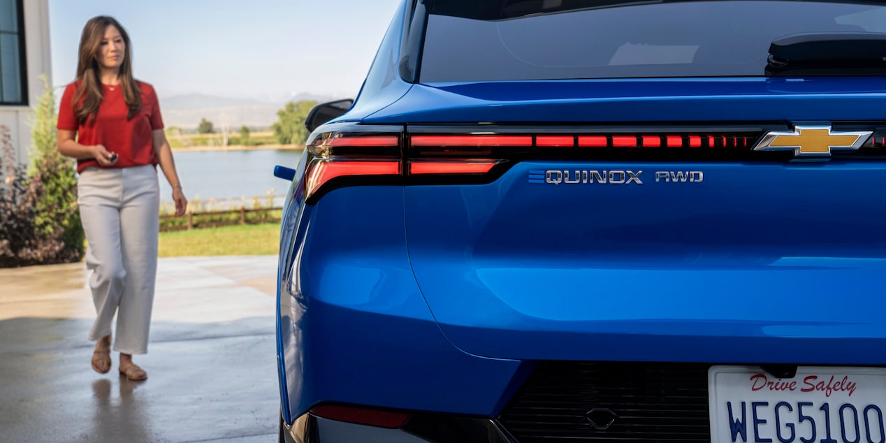 Rear View of a Blue 2025 Chevy Equinox EV to the Side of the Frame and a Woman Watching the Car