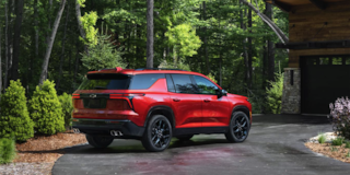 Three Quarters View of a Red 2025 Chevy Traverse in Front of a Wooden House Surrounded by Trees