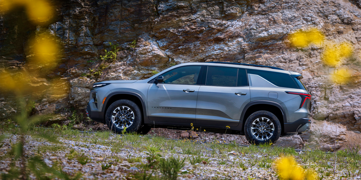 Side View of a Silver 2025 Chevy Traverse Driving Down a Road with a Mountain Wall to the Side