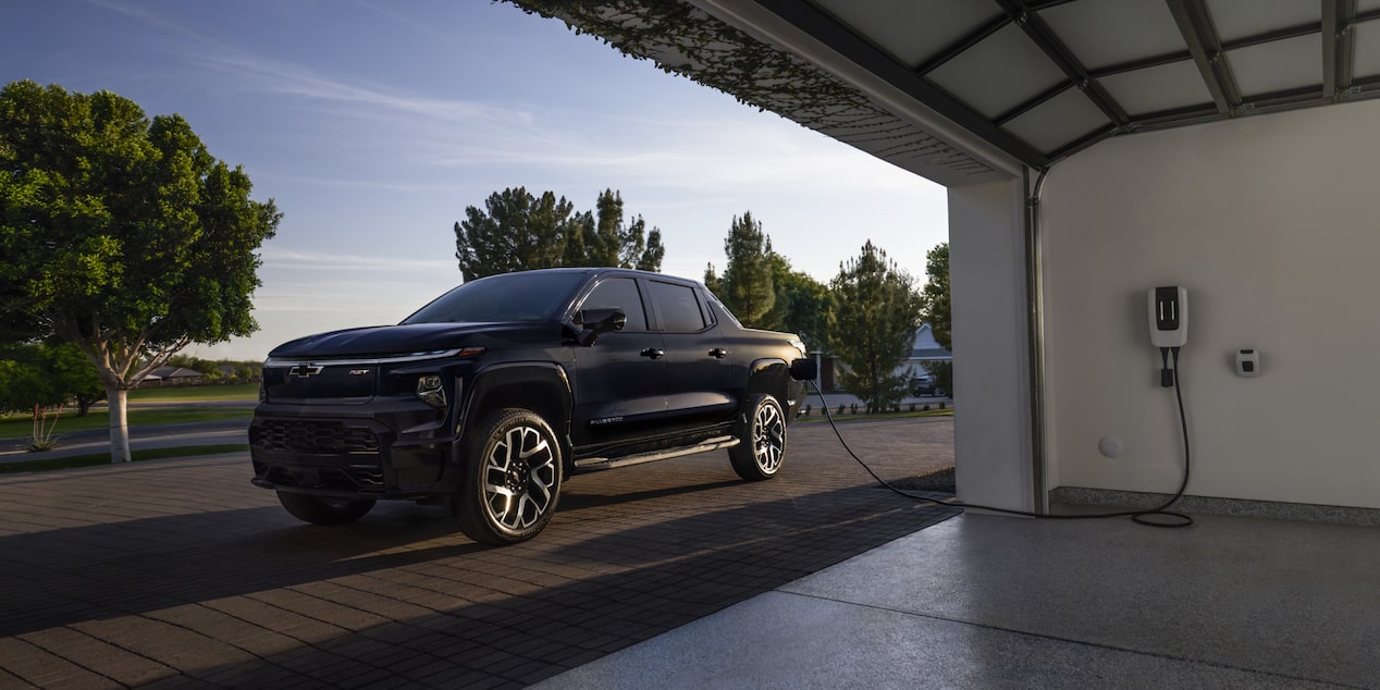 A black 2025 Silverado EV next to a garage getting charged with sunlight coming from the rear