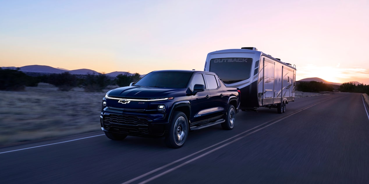 Three quarter view of a black 2025 Chevy Silverado EV with a trailer behind on a road at evening