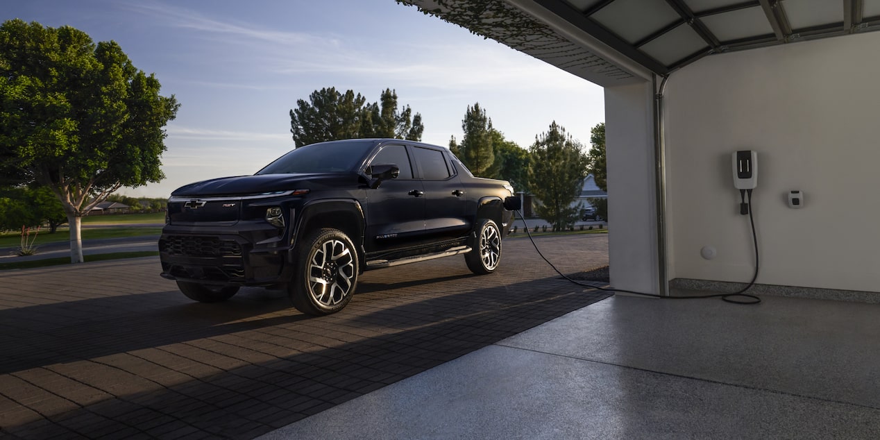 A black 2025 Silverado EV next to a garage getting charged with sunlight coming from the rear