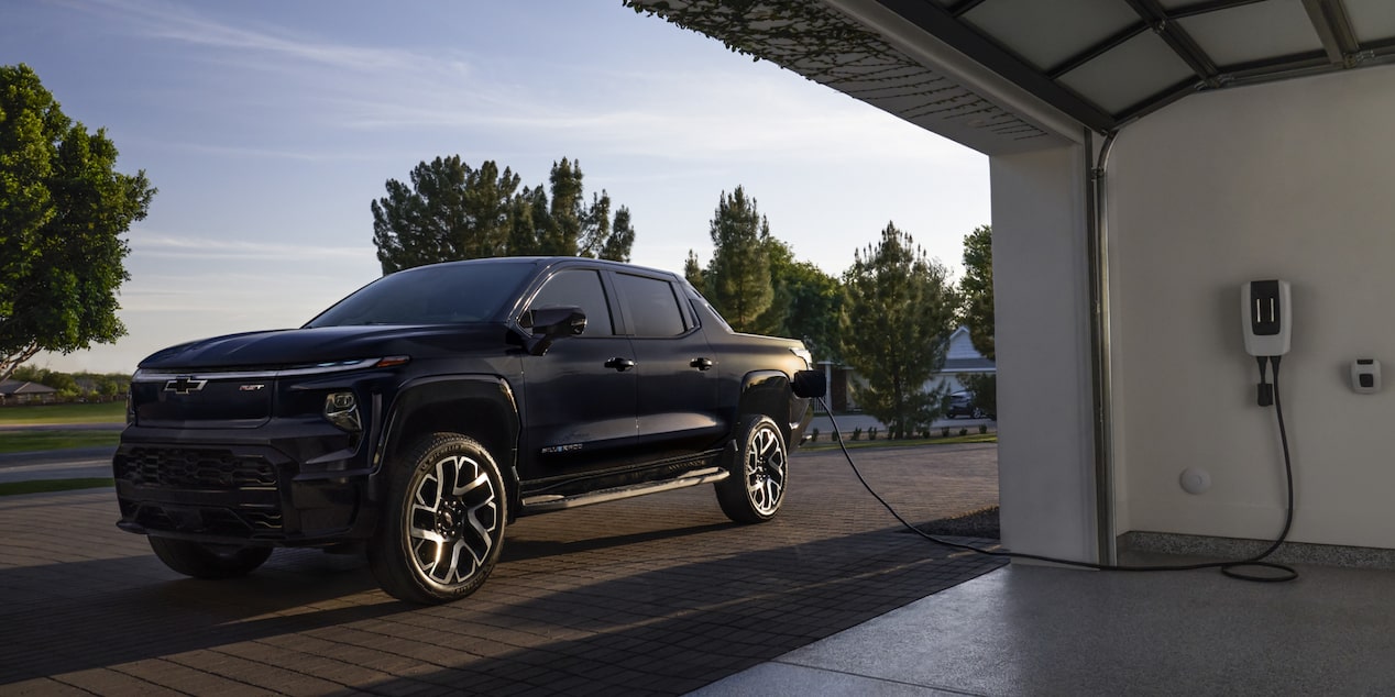 A black 2025 Silverado EV next to a garage getting charged with sunlight coming from the rear