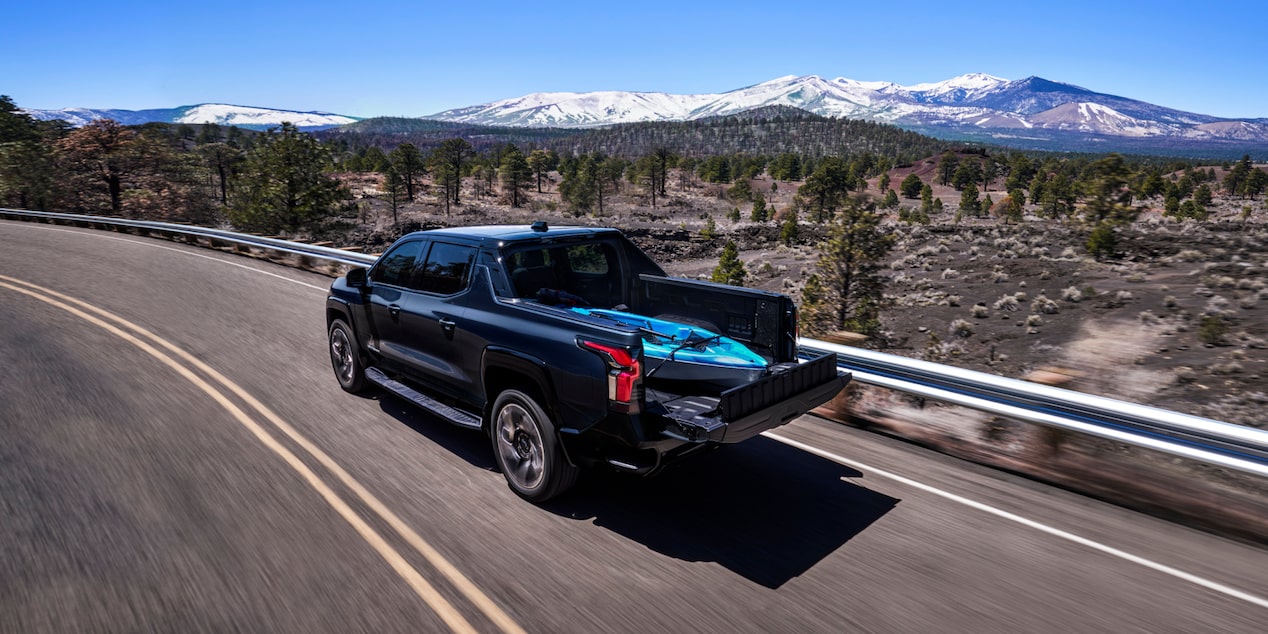 Rear three quarters view of a Chevy 2025 Silverado EV driving on the road next to mountains