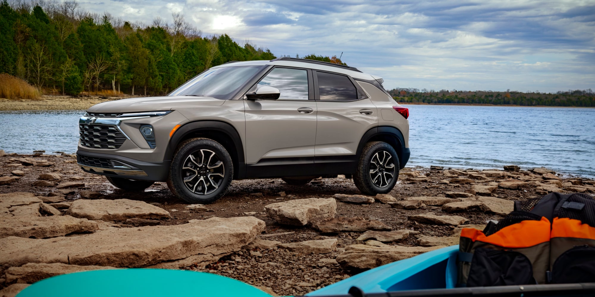 A Chevrolet Trailblazer Parked on a Rocky Lakeside Shore with Forest Trees and Calm Water Behind It.