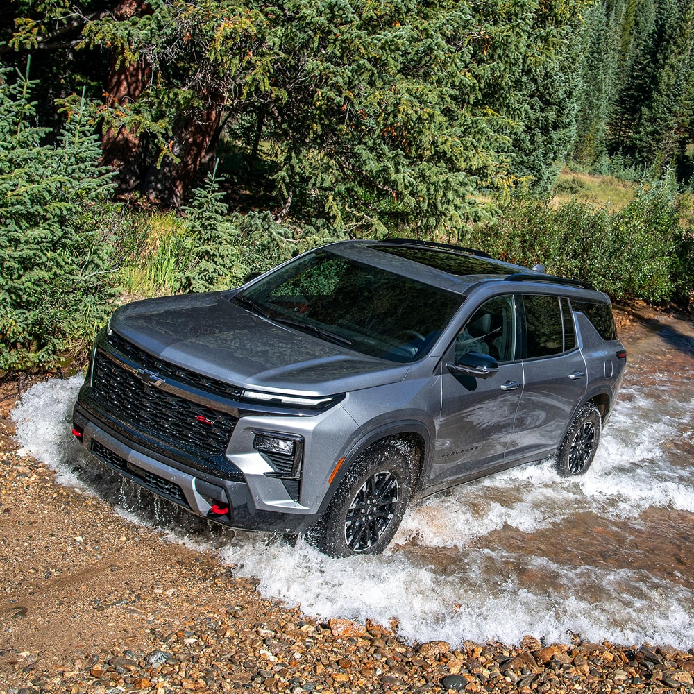 A 2026 Chevrolet Traverse Driving Through a Shallow Rocky Stream Surrounded by Dense Evergreen Trees.