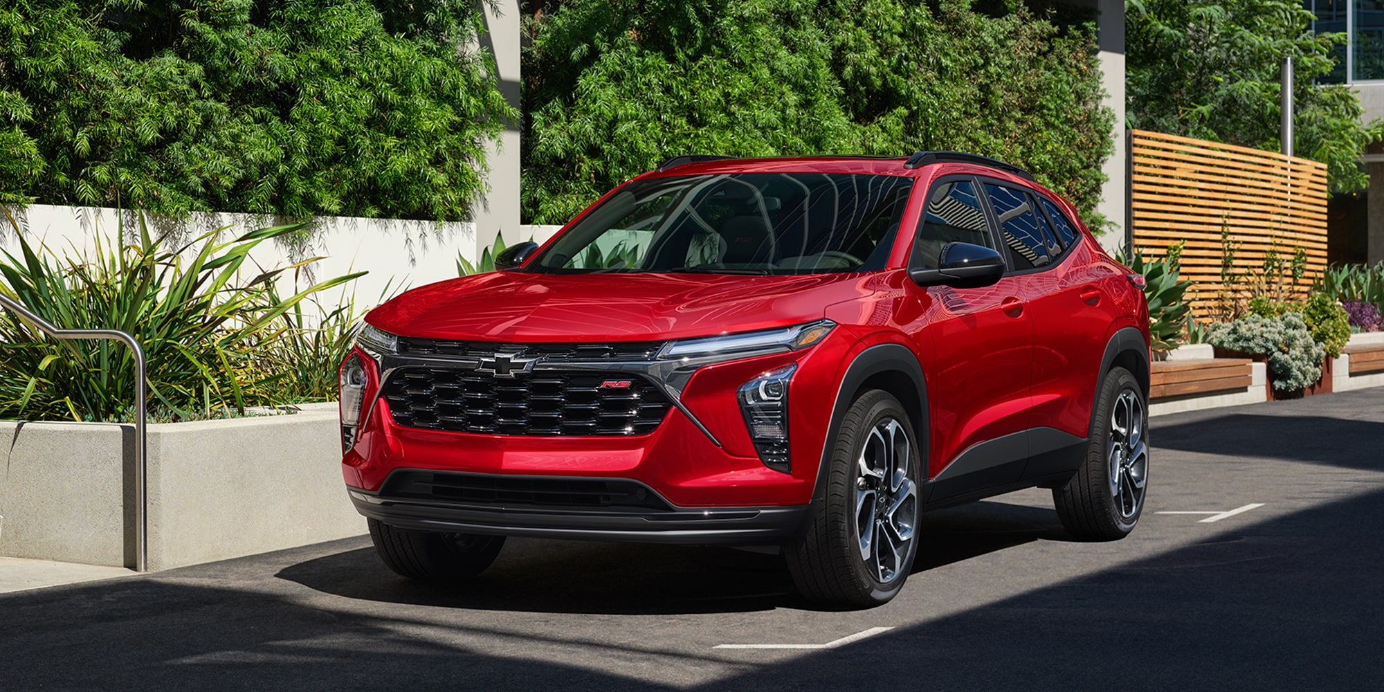 A Red Chevrolet Trax Parked in Front of a Modern Glass Building With Architectural Shadows Falling Across the Pavement.