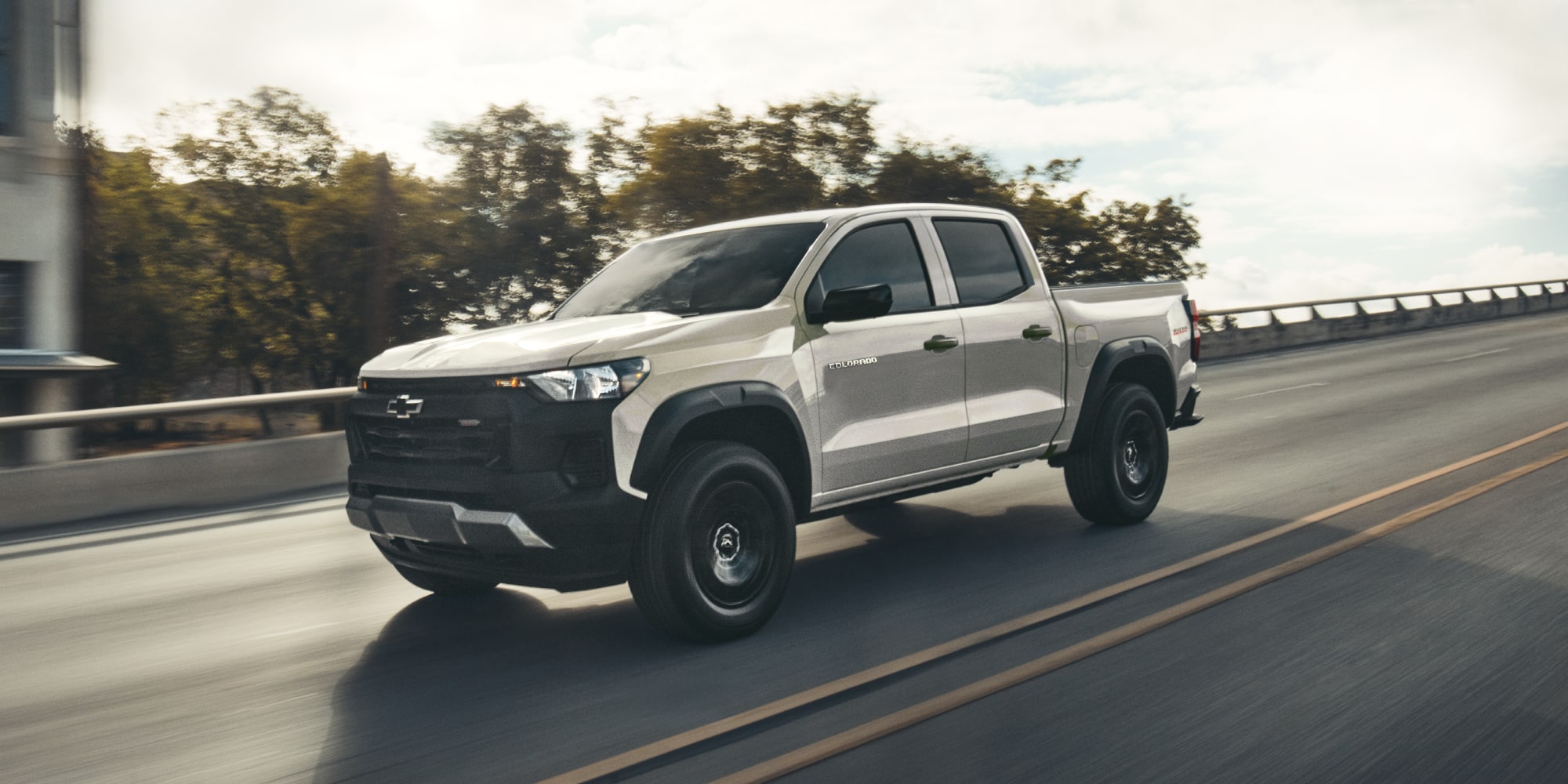 A 2026 Colorado Trail Boss in White Sands equipped with StowFlex Tailgate driving down the highway.