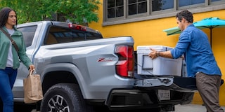 Three Quarters Rear View of a 2026 Silver Chevy Colorado Pickup Truck with a Man Putting a Box in the Trunk and a Woman Coming Towards Him