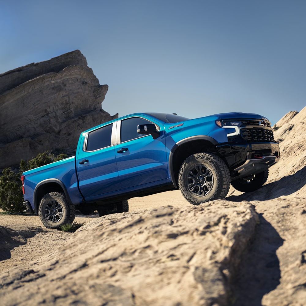 A Blue 2026 Chevrolet Silverado Driving Over Large Rock Formations in a Desert Landscape.