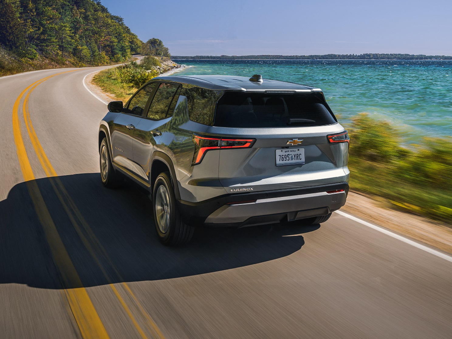 Rear View of a Chevy Equinox Driving Down a Road Outlooking the Ocean with Text Overlay Reading "Standard Safety and Driver Assistance Features"