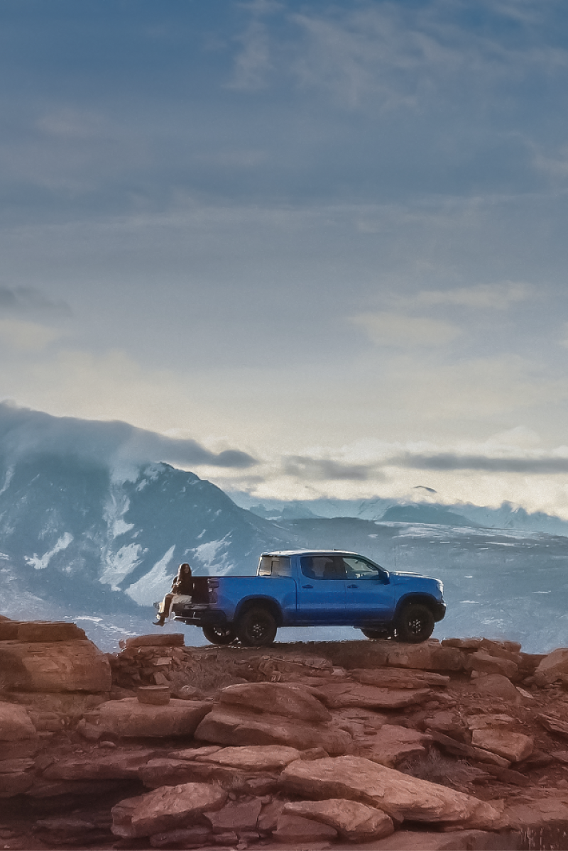A Blue Chevrolet Truck Parked on the Edge of a Massive Rock Cliff Overlooking a Mountain Range at Sunset.