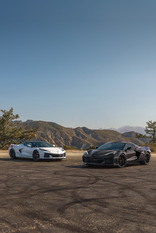 Two Chevrolet Corvette Grand Sport Sports Cars, One White and One Black, Parked on a Sunlit Mountain Overlook with Rugged Hills and Sparse Trees in the Distance.
