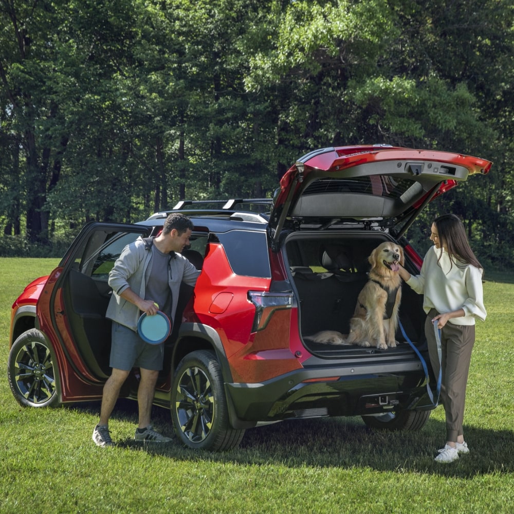 Red Chevrolet SUV Parked on a Grassy Field With the Rear Liftgate Open and a Golden Retriever Sitting in the Cargo Area While Two People Stand Nearby.