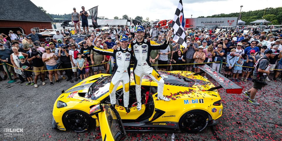 Corvette Racing Drivers Standing on Vehicle Holding up Racing Flag and Smiling with an Audience in the Background