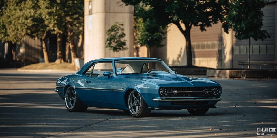View of a Blue 1968 Chevrolet Camaro Covered by Shade Parked in Front of a Building 