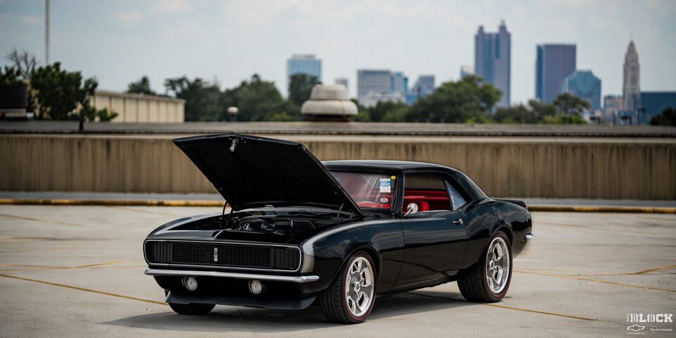 Three Quarters View of a Black 1968 Chevy Camaro Parked in an Empty Lot with its Hood Open and Skyscrapers in the Distance