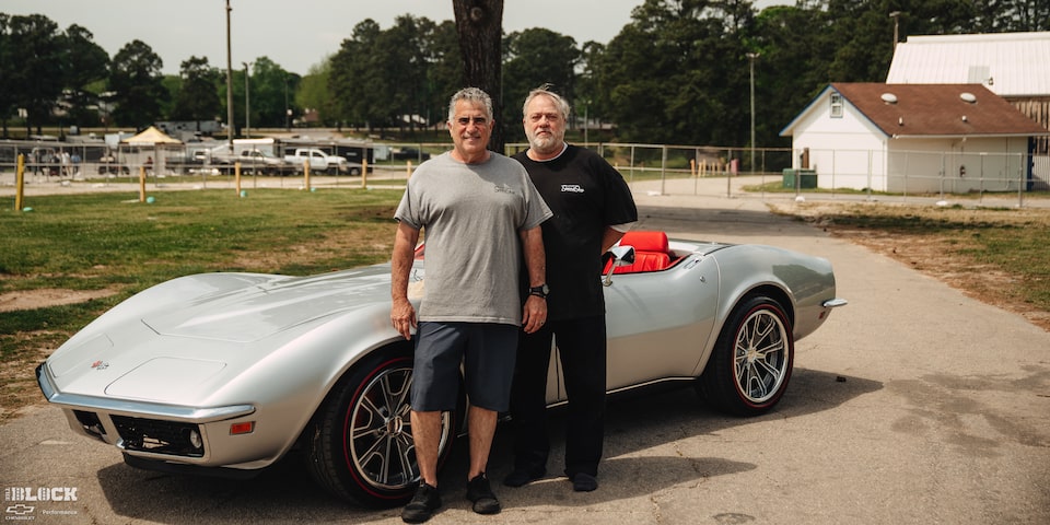 Two Men Standing in Front of a 1969 Silver Chevy Corvette with a Tree in the Back along with a House in the Distance