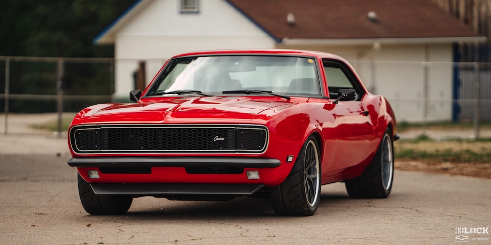 Three Quarters View of a 1968 Orange Chevy Camaro on a Road with a House Behind It
