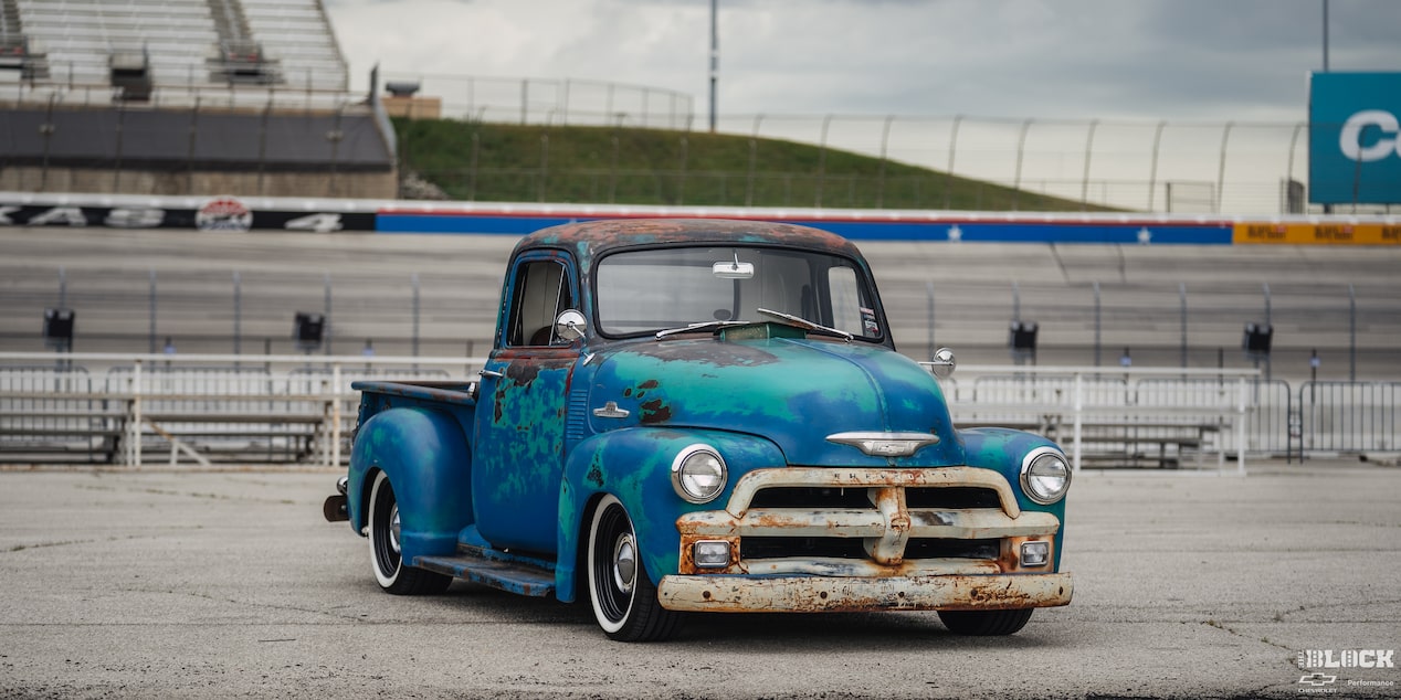 Vista de medio perfil de una pickup Chevy 1955 oxidada al costado de una pista de carreras