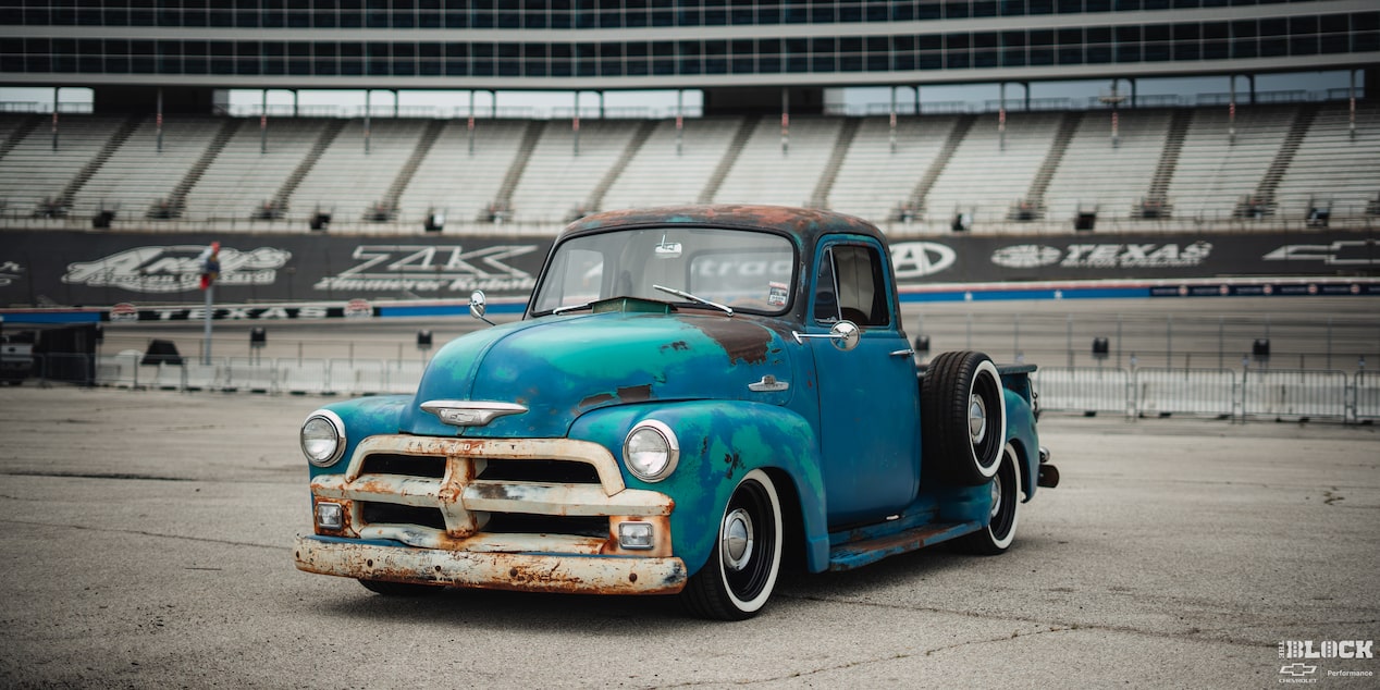 Vista de medio perfil de una oxidada pickup Chevy 1955 en Azul, en frente de un estadio de carreras