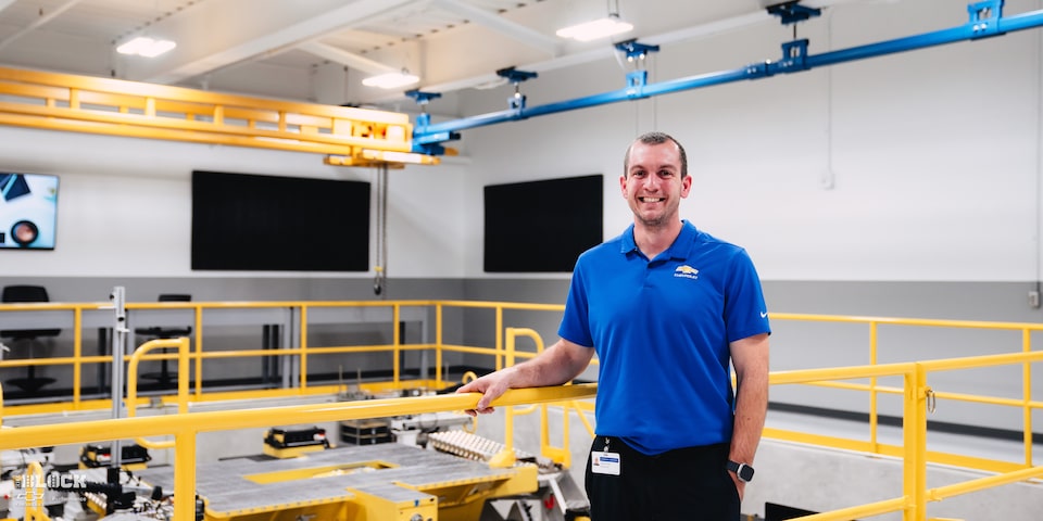 A GM Employee in a Blue Chevrolet Shirt Smiles and Stands In Front of Machinery at the GM Charlotte Technical Center.