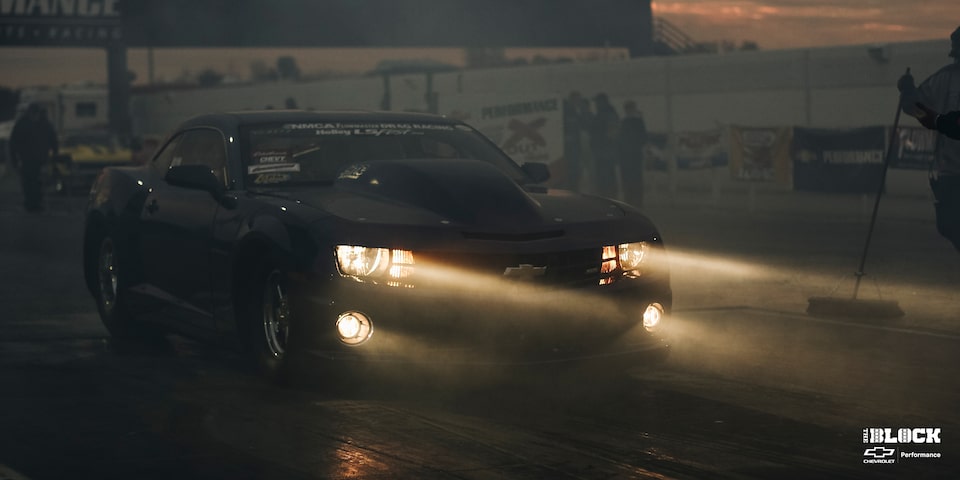 A Chevrolet Copo Camaro Driving on a Race Track at Night With Its Headlights On