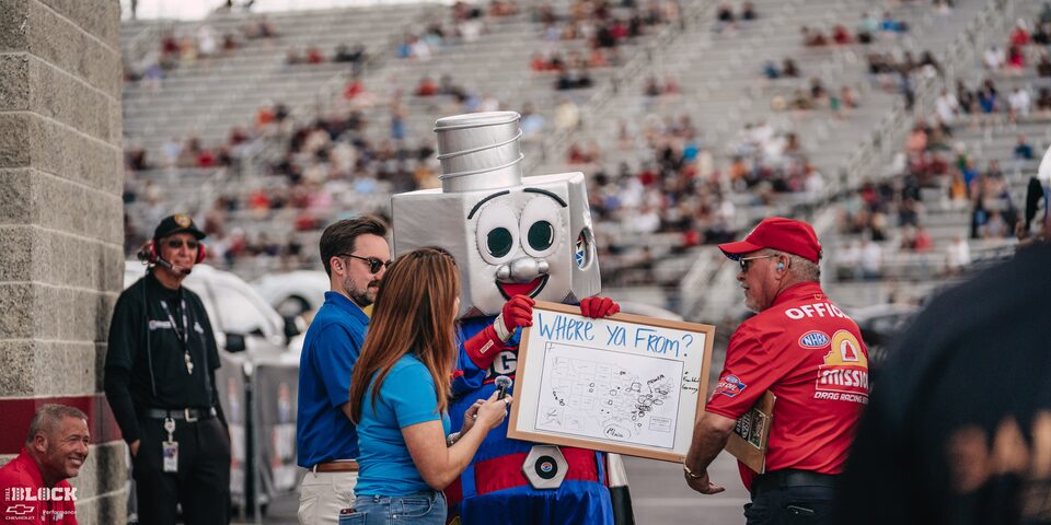 Attendees Interact with a Mascot in a Smiling Lugnut Costume Holding a Whiteboard With a Map of the United States and Text Reading "Where Ya From?" at Charlotte Motor Speedway