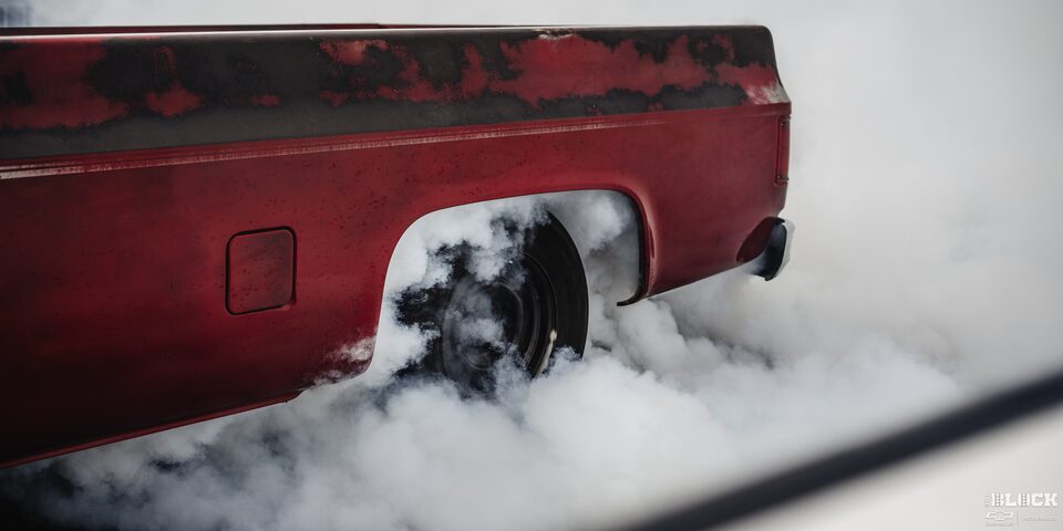 Close-up of a the Rear Left Wheel of a Cherry Red Chevy Truck With Black Accents as it Burns Rubber Spewing Smoke